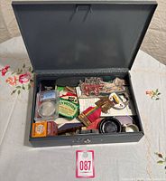 Metal cashbox open showing various small keepsakes inside including coins, pins, patches, and cards.