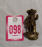 Vintage brass Prince Charlie miniature door knocker positioned next to a lot number card showing size and general appearance of the item.