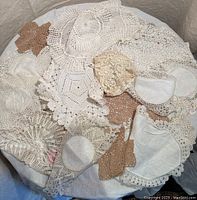Top view of assorted vintage doilies in white, cream, beige, and brown hues, showing intricate crochet and lace patterns of various shapes including round, hexagonal, and floral.