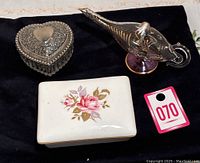 Front view of the three items on black fabric: heart-shaped jewellery dish, floral trinket box, and decorative lantern shaped like a genie lamp