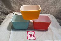 Three vintage Pyrex refrigerator dishes each in pastel colors stacked with yellow on top, light blue and reddish-orange below.