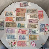 Top-down view of first set of vintage Yugoslavian paper money spread on a round floral tablecloth showing various denominations with portraits and architectural imagery.