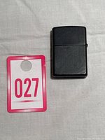 Closed vintage Zippo lighter with matte black finish sitting on a white surface next to a red and white numbered tag '027'.
