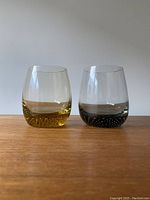 Front view of two oval shaped drink glasses with colored bases, yellow and black, on wooden surface against plain wall