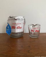 Three new Anchor Hocking Fire-King glass measuring cups, clearly showing two of them with red logos and measurement markings on a wooden surface against a plain wall.