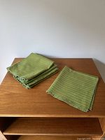 Six green textured fabric place mats folded and stacked on a wood surface.
