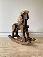 Front side view of the wooden rocking horse showing wool mane and hand grip handles on the head. The toy stands on curved rockers and has a saddle strap around the top of the back.