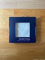 Front view of unopened bas relief painting in dark blue packaging with a small transparent window showing the white textured artwork with birds and geometric shapes.