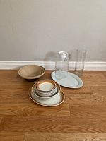 Photo showing entire kitchenware set including bowls, plates, glass pitcher, tall glass, and serving platter on wooden floor against white wall.