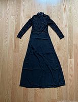 Full view of long black dress laid flat on a wooden floor showing long lace sleeves, high neck, and long skirt.