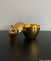 Gold-tone metal apple trinket box with lid removed showing hollow interior, sitting on table with neutral background