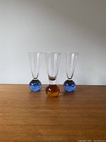 Front view of the set of three shot glasses on a wooden surface against a plain wall, showing two blue base glasses flanking one amber base glass.