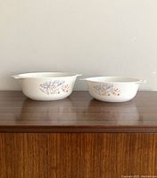 Two vintage Pyrex milk glass bowls with floral designs side by side on a wooden surface, showing their exterior and design.