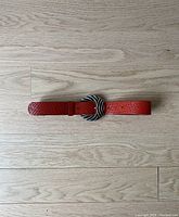 Red textured leather belt with metal buckle laid flat on wooden floor