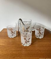 Photo showing full set of frosted glass ice bucket and five drinking glasses with gold leaf pattern arranged on wood surface.