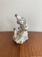 Front and side view of the porcelain Pierrot clown figurine playing a concertina, seated on a blue stool with a dog on dice by its side.