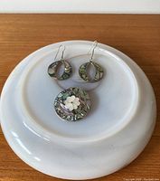 Top down photo of the brooch and earrings on white ceramic plate showing abalone shell colors and floral detail on brooch.