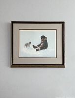 Framed vintage print depicting an Inuit boy sitting in the snow playing with two puppies. The frame is gold-toned with visible age wear.