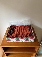 Box containing folded orange fabric place mats and hand towels along with wooden napkin rings, all arranged inside a decorative gift box with holiday-themed wrapping paper.