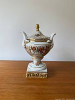 Front view of the vintage porcelain urn showing floral patterns, gold details, and finial on lid.