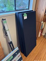 Two rectangular black fabric-covered bass trap panels standing upright against a wall by a window and hardwood floor.