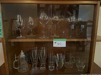 Wide view of wooden cabinet with two shelves of assorted glassware, including stemmed wine glasses, beer stein, tall patterned glasses, and small tumblers.