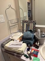 Full view of bathroom items including towels, mirror, toiletry bottles, towel rack, basket, box of adhesive bandages, and other personal care items on countertop.