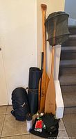 Photo showing two wooden paddles, rolled up black sleeping bag and camping mat, insect head net hat, water bottle, gloves, and small accessories grouped on floor next to stairs.