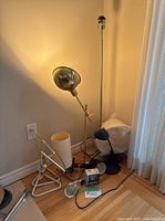Photo showing six assorted lamps in a corner, including a tall silver floor lamp, a white table lamp with a fabric shade, a black adjustable lamp, and some lamp parts with cords on the floor.