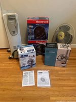 Photo showing the DeLonghi radiator heater, four boxed fans (Garrison, two Honeywell, Hampton Bay), and manuals on hardwood floor.
