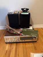 Mitsubishi DA-R8 stereo receiver on wooden floor with two Paradigm bookshelf speakers behind it, user manual atop receiver, and speaker wires laid out in front.