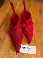 Pair of hand-felted red mouse slippers sitting on wooden floor, showing front and side mouse features including ears, eyes and tails with colored pom-poms.