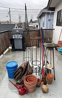 Photo of entire lot with grill background, includes terra cotta pots, plastic pots, metal plant stands, blue bucket, brooms, watering can, tarp.
