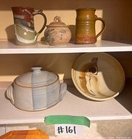 Five pottery pieces on two cabinet shelves: two handled mugs in different glazes, a small round jar with lid and animal figure, a large covered casserole dish, and a bowl.