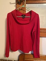 Red cashmere sweater hanging on a green hanger in front of wooden doors, showing overall view of the sweater with long sleeves and cowl neckline.