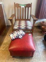 Front view of the vintage solid oak chair with reupholstered geometric patterned seat, red leather-like ottoman, and folded extra fabric.
