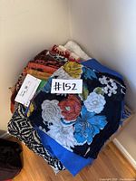 Stack of assorted scarves including a floral printed scarf on top, folded and stacked against wall on floor.