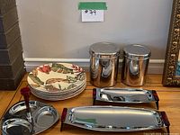 View of 6 decorative ceramic plates with tropical leaf and pineapple motifs stacked, two stainless steel canisters with lids, a divided stainless steel serving plate with three compartments and a central red handle carousel, and two stainless steel rectangular serving trays with red handles and foldable legs on a wood surface.