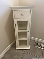 Full frontal photo of white bathroom cabinet showing drawer and glass-door with shelves inside