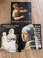 Three hardcover art books placed on wooden floor showing front covers: Masters of Venice, American Folk Painting, Dutch Painting of the Golden Age