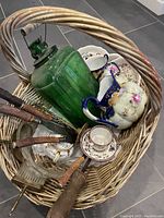 Photo showing woven basket with large green antique glass bottle, stack of floral pattern dishes, Royal Nippon cup and saucer, carving set with wooden handles, brass silent butler, and pressed glass items.