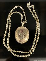 Front view of the antique oval locket pendant hanging on the rope chain, showing the etched border and floral motif with pearls and red stones.