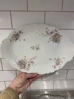 Hand-held view of the oval porcelain platter showing purple and green floral decorations, embossed ornate rim, and overall condition without chips or cracks.