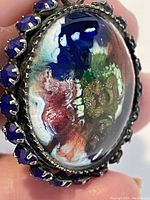 Close-up of a vintage brooch or pin with a colorful cameo scene, set in a silver-tone metal frame decorated with blue beads around the edges.
