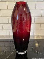 Front view of the sleek ruby red glass vase against a white tiled background, showing smooth glossy surface and gradation of color from dark to red.