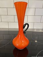 Side profile of the orange art glass vase with black handle showing slender neck and wide base.