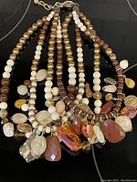 Close-up of necklace showing variety of beads in brown, white, cream, amber, and metallic tones with faceted and smooth finishes, including large teardrop-shaped glass beads and coin-like metal charms.