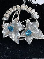 Sterling silver pendant or pin shaped as two textured leaves with round blue stones and a semi-circle of baguette-cut clear stones above.