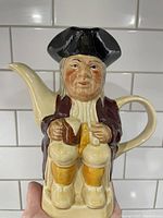 Front view of a hand-painted ceramic teapot in the shape of a seated figure wearing a tricorn hat, maroon coat, and yellow stockings.