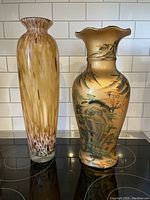 Two large decorative vases side by side on a kitchen counter with white subway tile background. Left vase is tall art glass with gold flecks and multicolored swirls. Right vase is oriental style with painted natural landscape design and ruffled top.